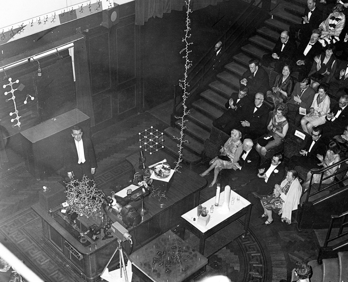 Just months after the X-ray structure of lysozyme was published in 1965, David C. Phillips gave a public lecture in London. In this photo, on the table in front of him is a folded model of lysozyme. Hanging from the ceiling is the unraveled version, with all 129 amino acids represented.