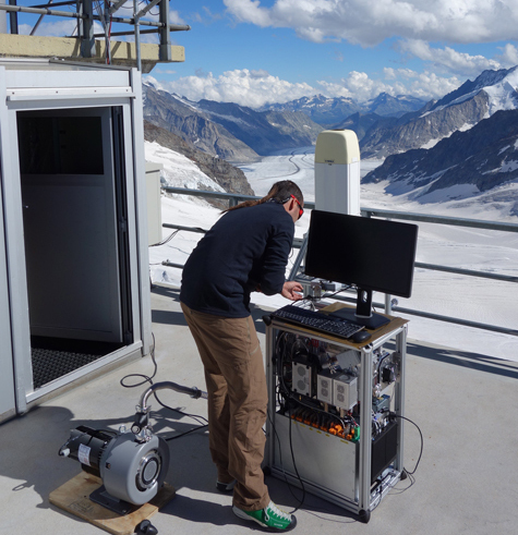 A man, standing with his back to us, is leaning over some equipment. He is standing on a balcony with a view over a glacier.