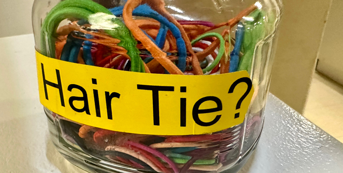 A jar filled with hair ties. On the jar is a sticker saying “Need a hair tie?”