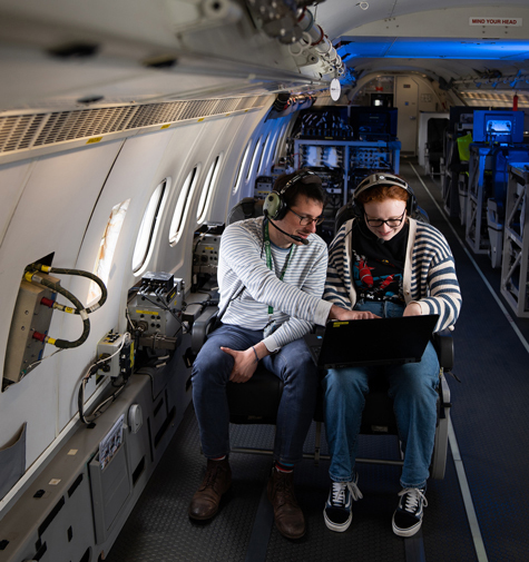 Two people sit in an airplane looking at a laptop screen. Behind them are racks of equipment instead of more seats.