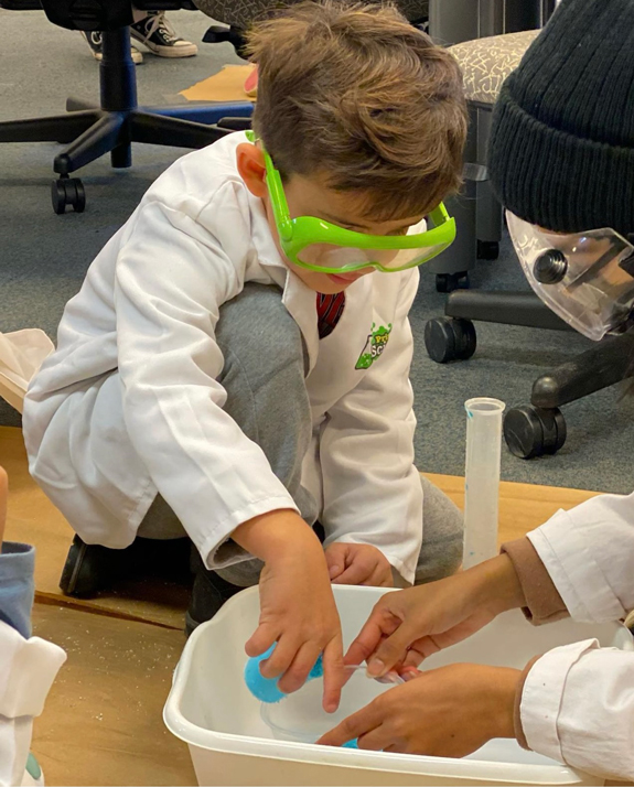 A small child in safety goggles and lab coat kneels on the ground and puts a hand in a large tray as he’s helped to do an experiment.