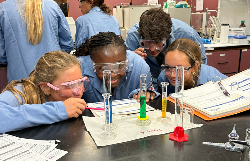 Students in lab coats and safety goggles kneel down to have their eyes at the same level as the colored liquids in measuring cylinders that are lined up on the bench.