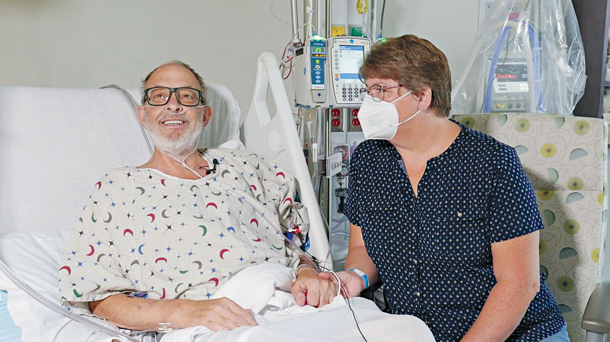 Lawrence Faucette smiles while lying in a hospital bed. He holds the hand of his wife, Ann. Ann is sitting in a chair next to the bed wearing a respirator mask.